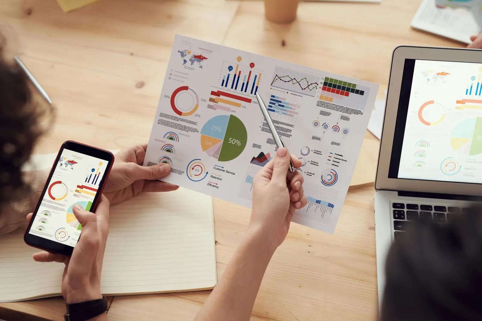 Hands holding a phone and pen over printed media charts beside a laptop
