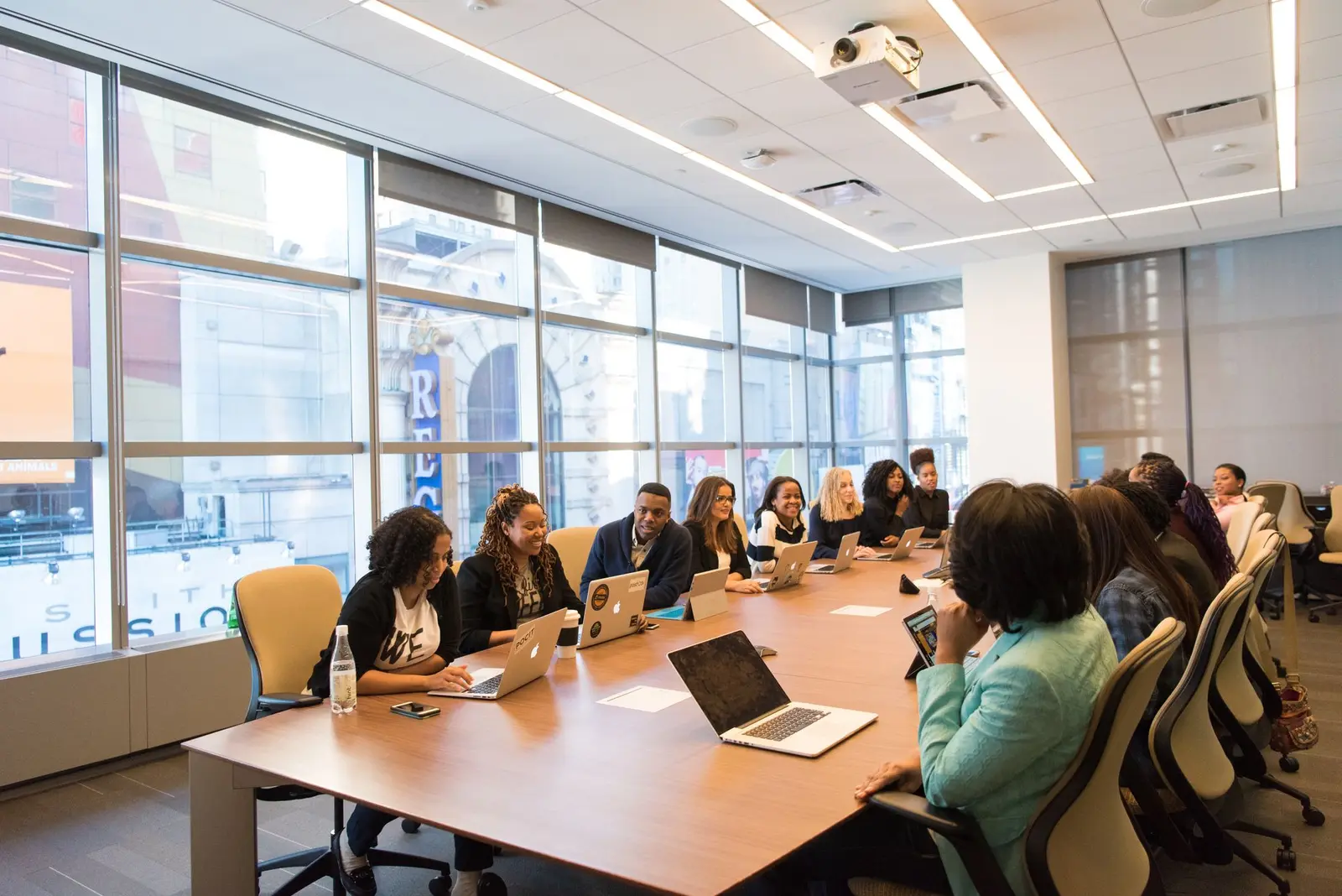 A bright boardroom with a long table where a group listens to a presenter at a whiteboard