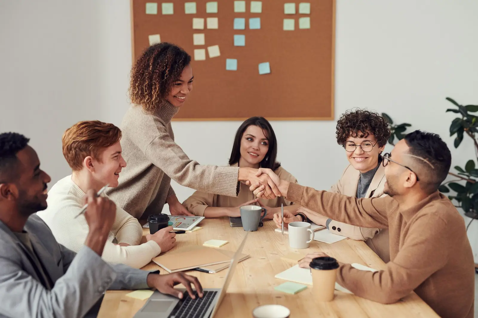 Colleagues shaking hands across a meeting table in a bright office with a pinboard of notes behind them